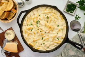 A skillet filled with alfredo pasta next to garlic bread and grated parmesan cheese.
