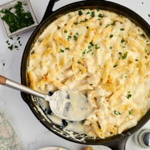 Skillet of cheesy baked penne pasta and alfredo being served, with parsley garnish nearby.