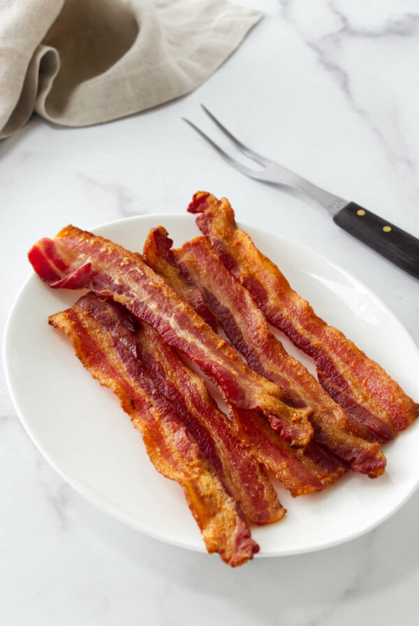 Six crispy bacon strips on a white oval plate, fork resting nearby.