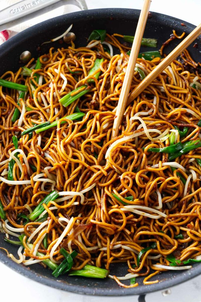 Pan fried noodles in a skillet with scallions and chop sticks.