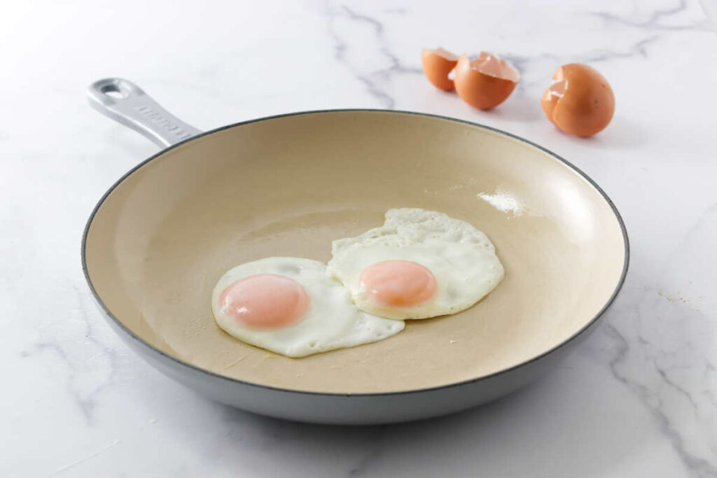 Two fired eggs in a white skillet in front of egg shells.