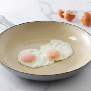 Two fired eggs in a white skillet in front of egg shells.