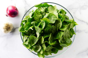 A bowl of baby spinach leaves next to a red onion.