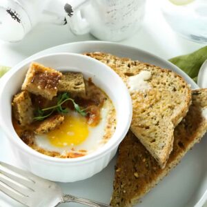 Baked egg in ramekin with croutons, herbs, and toast on white plate.