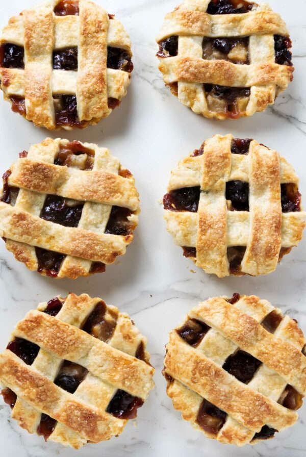 Six mini lattice-topped dried cherry tarts with golden crusts on white marble.