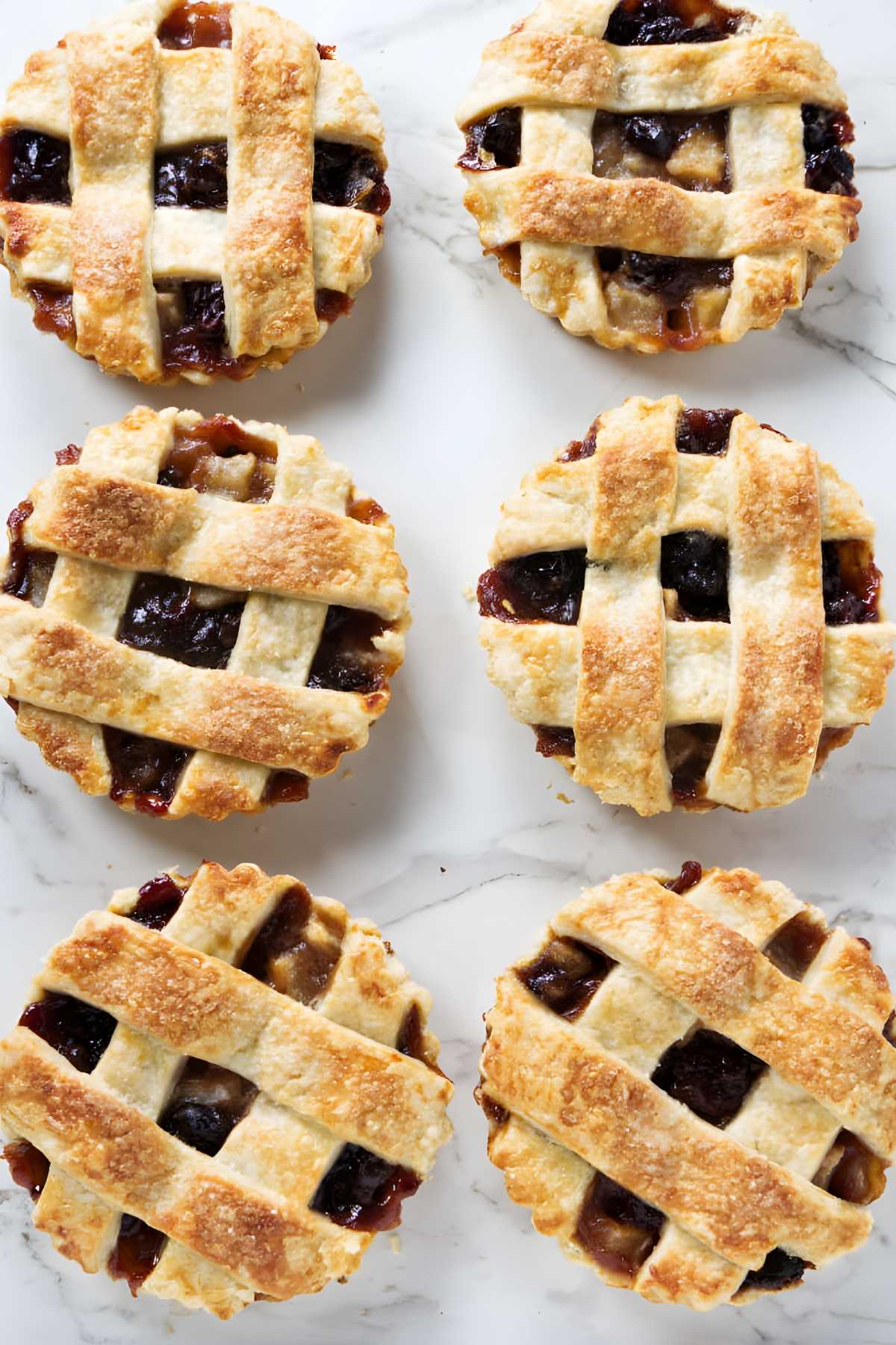 Six mini lattice-topped dried cherry tarts with golden crusts on white marble.