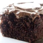 Chocolate cake slice with glossy frosting on a white plate and fork.