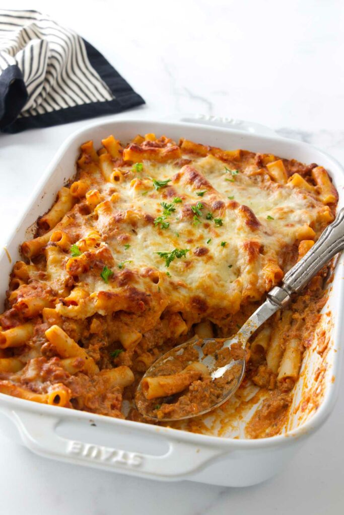 A serving spoon scooping ziti pasta from a baking dish.