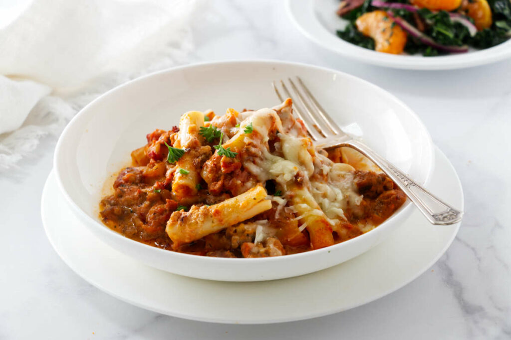 Bowl of cheesy baked pasta with parsley and fork, salad in background.