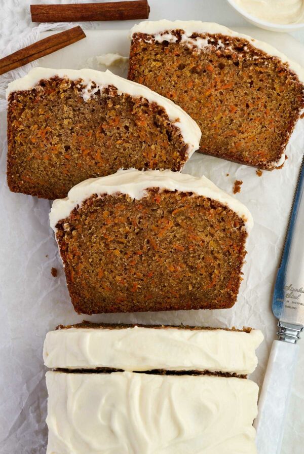 Sliced carrot cake quick bread with sour cream frosting on parchment, knife and cinnamon nearby.