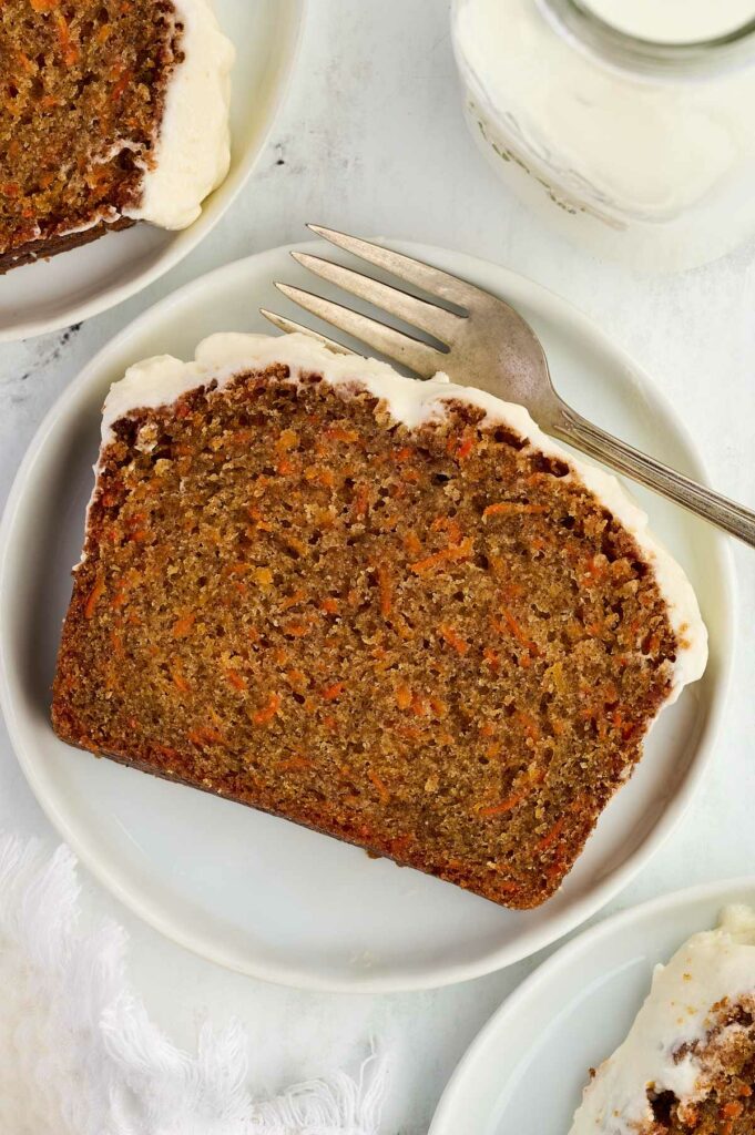 Carrot cake bread slice with cream cheese frosting on plate, fork, and milk.