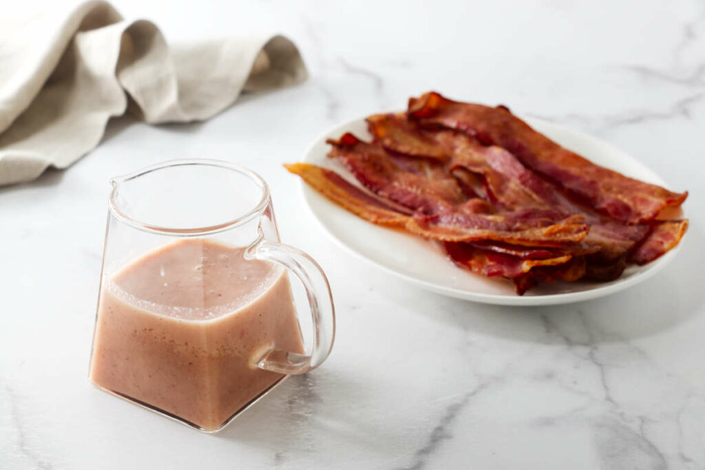 A glass pitcher filled with hot bacon dressing next to a plate of bacon.