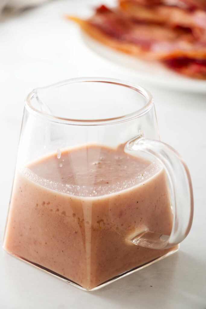 A clear glass jar filled with warm bacon dressing in front of a pile of crisp bacon.