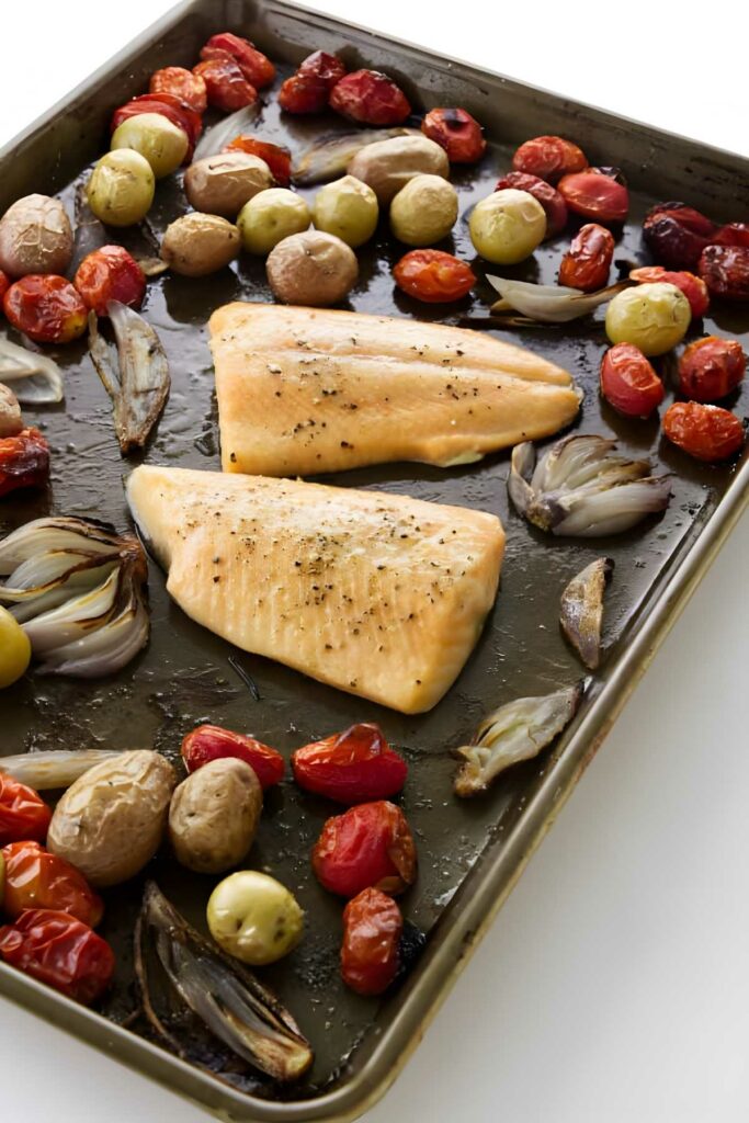 Two seasoned arctic char fillets with roasted potatoes and vegetables on a baking sheet.