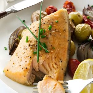 Cooked salmon with chives, roasted vegetables, and lemon on a plate; fork holding bite.