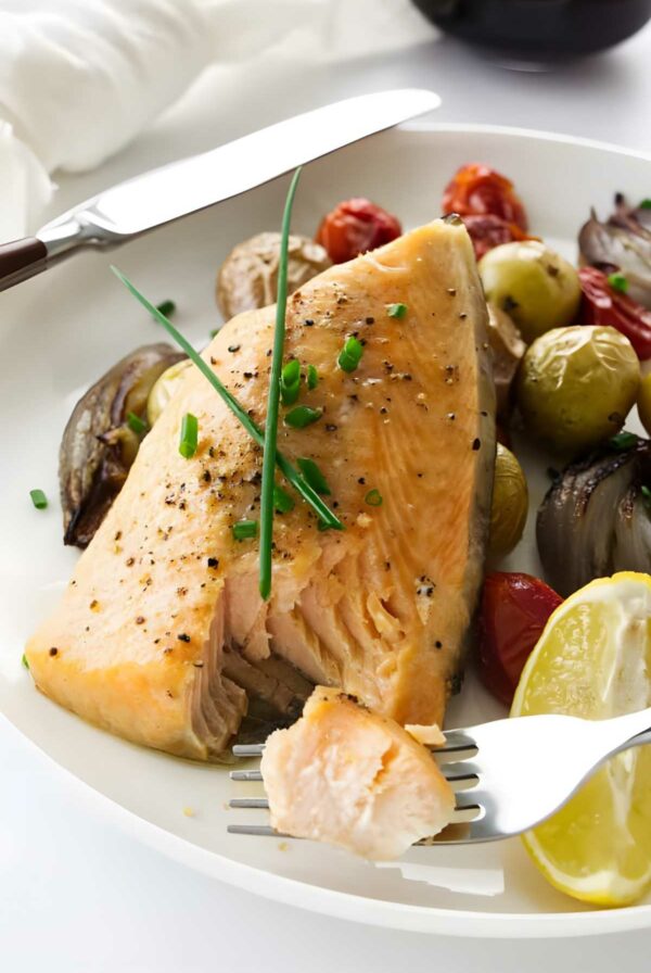 Baked arctic char with chives, roasted vegetables, and lemon on a plate; fork holding bite.