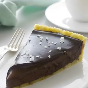Chocolate tart slice with yellow crust and sea salt on a white plate.