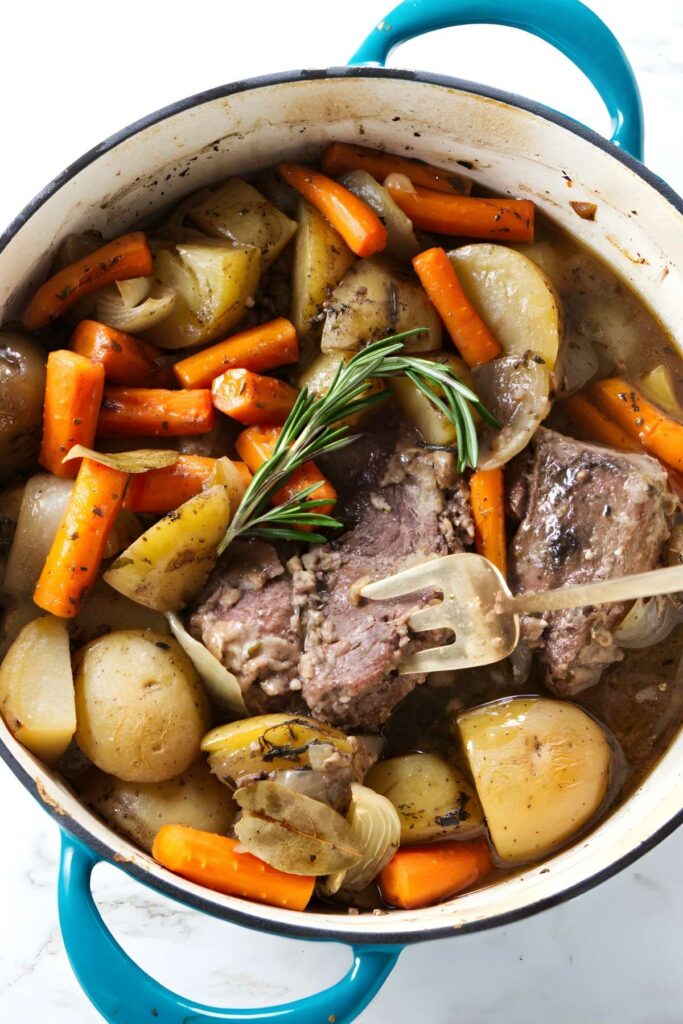 Blue pot of pot roast with beef, potatoes, carrots, herbs, and fork.