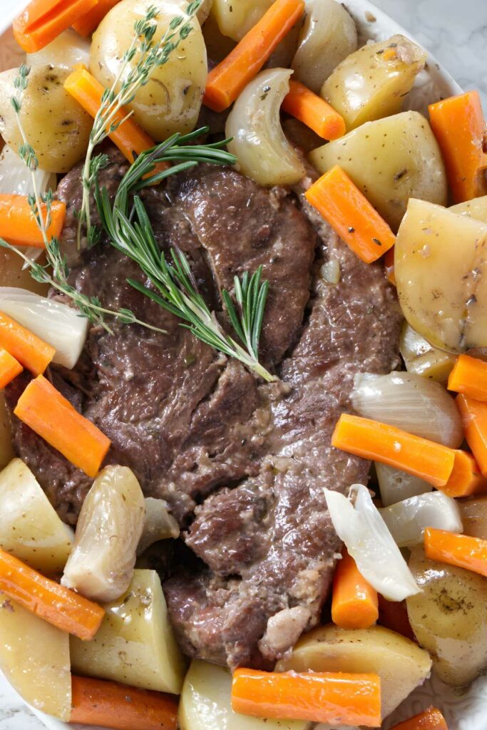 Pot roast with rosemary, thyme, potatoes, carrots, and onions on white plate.