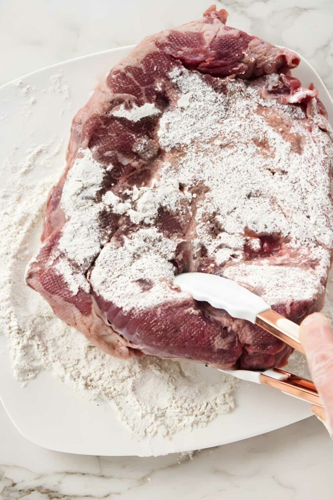 Hand with tongs coats raw meat in flour on a white plate.