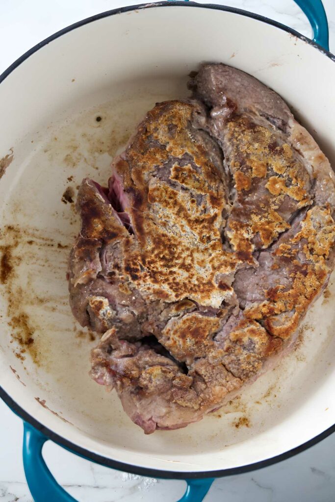 Browned meat searing in white pot with blue handles, partial crust visible.