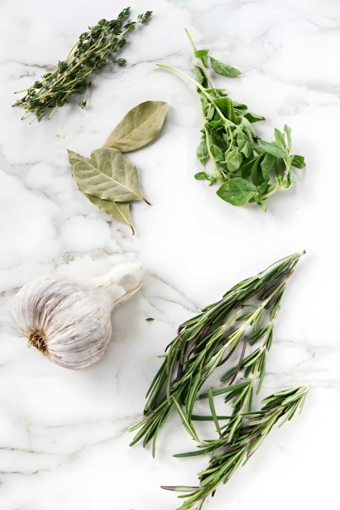 Assorted fresh herbs and seasonings on white marble, with garlic bulb.