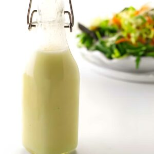 Glass bottle of creamy salad dressing on white surface, salad blurred behind.