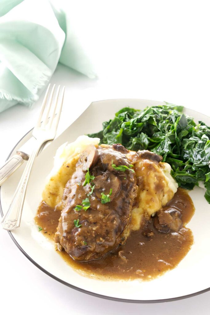 Salisbury steak with gravy, mashed potatoes, greens on a plate with utensils.