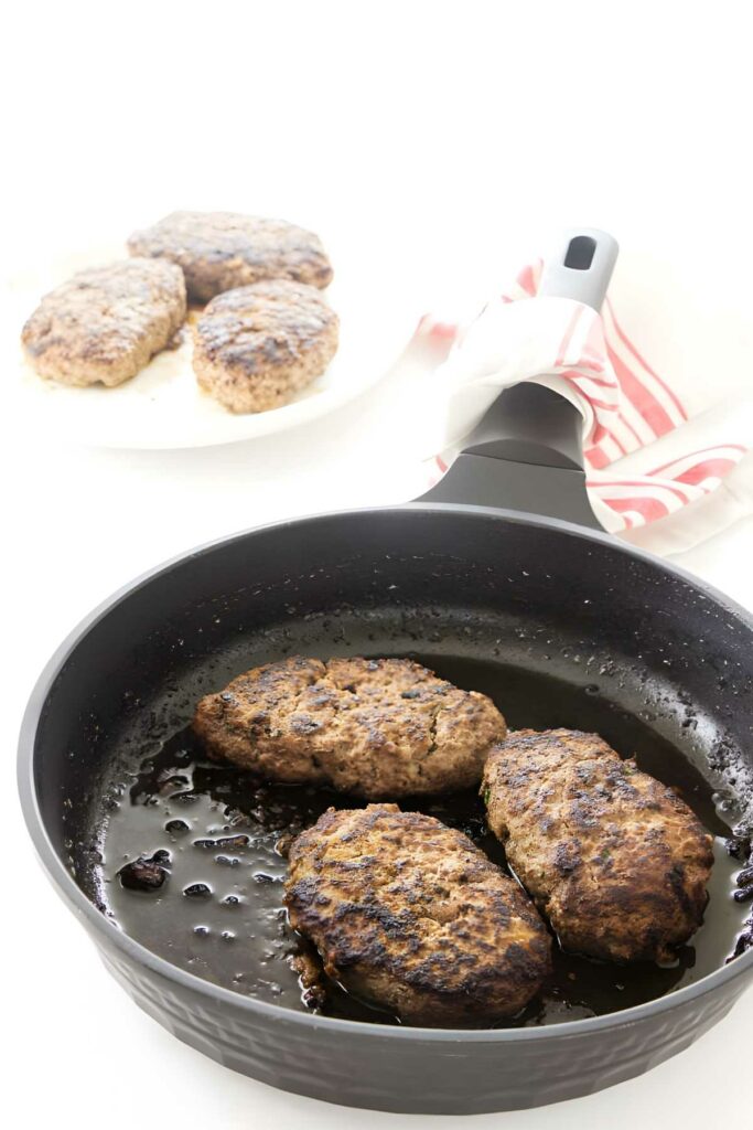 Three cooked hamburger patties in a black pan, more patties on plate behind.