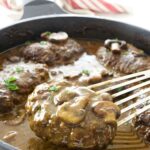 Salisbury steaks with gravy and mushrooms in a skillet; spatula lifting one steak.
