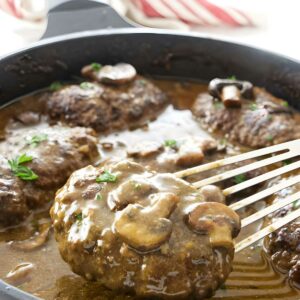 Salisbury steaks with gravy and mushrooms in a skillet; spatula lifting one steak.