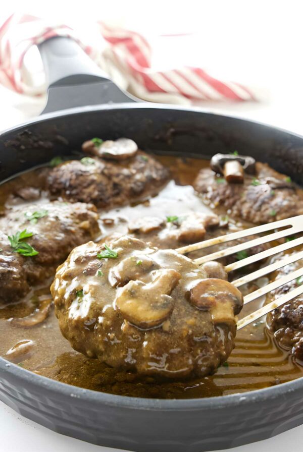 Salisbury steaks with gravy and mushrooms in a skillet; spatula lifting one steak.