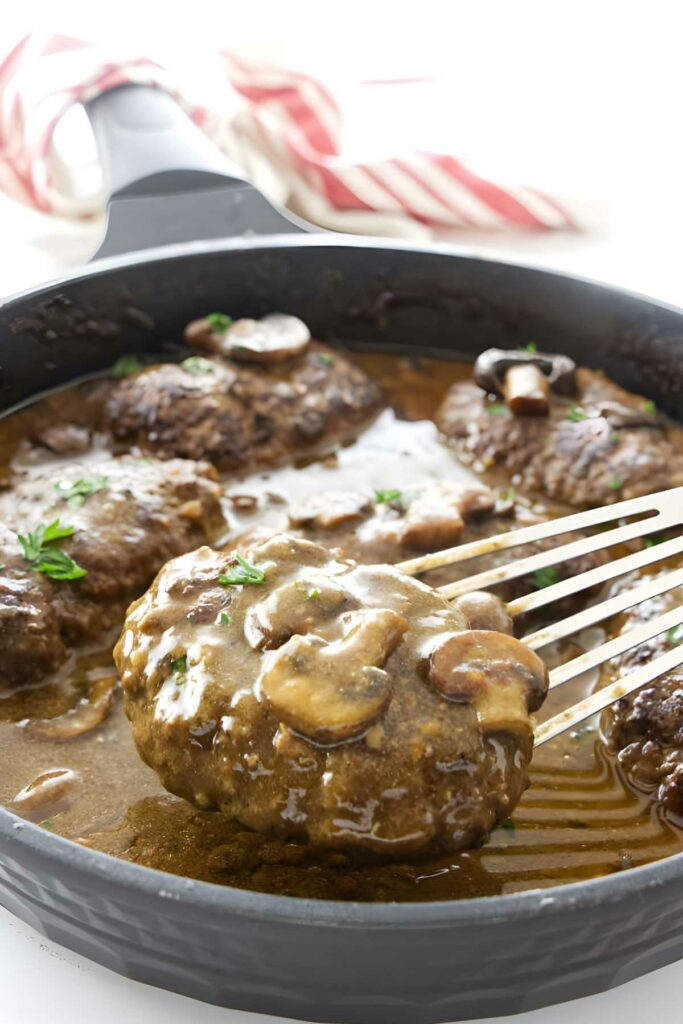 Salisbury steaks with gravy and mushrooms in a skillet; spatula lifting one steak.