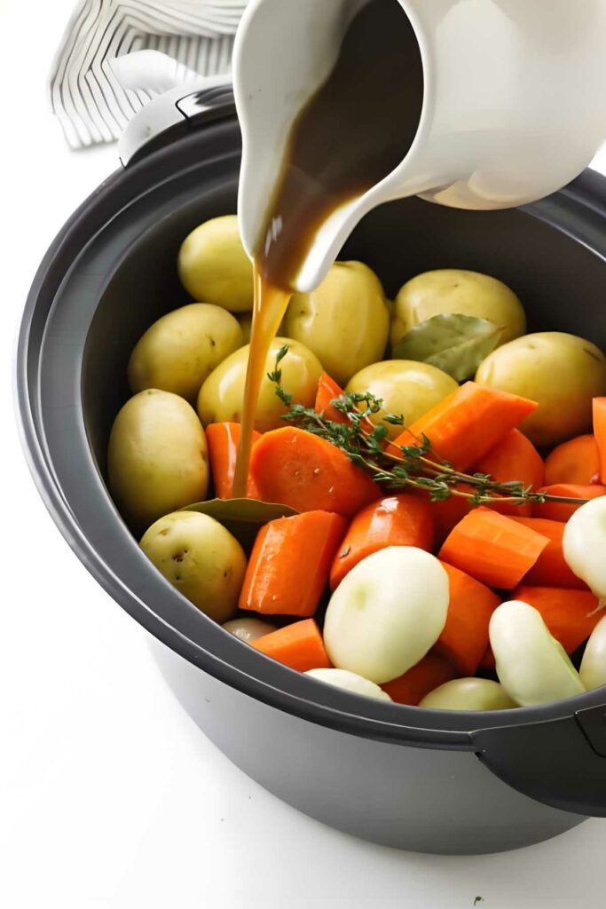 Broth is poured from a jug over vegetables and herbs in a slow cooker.