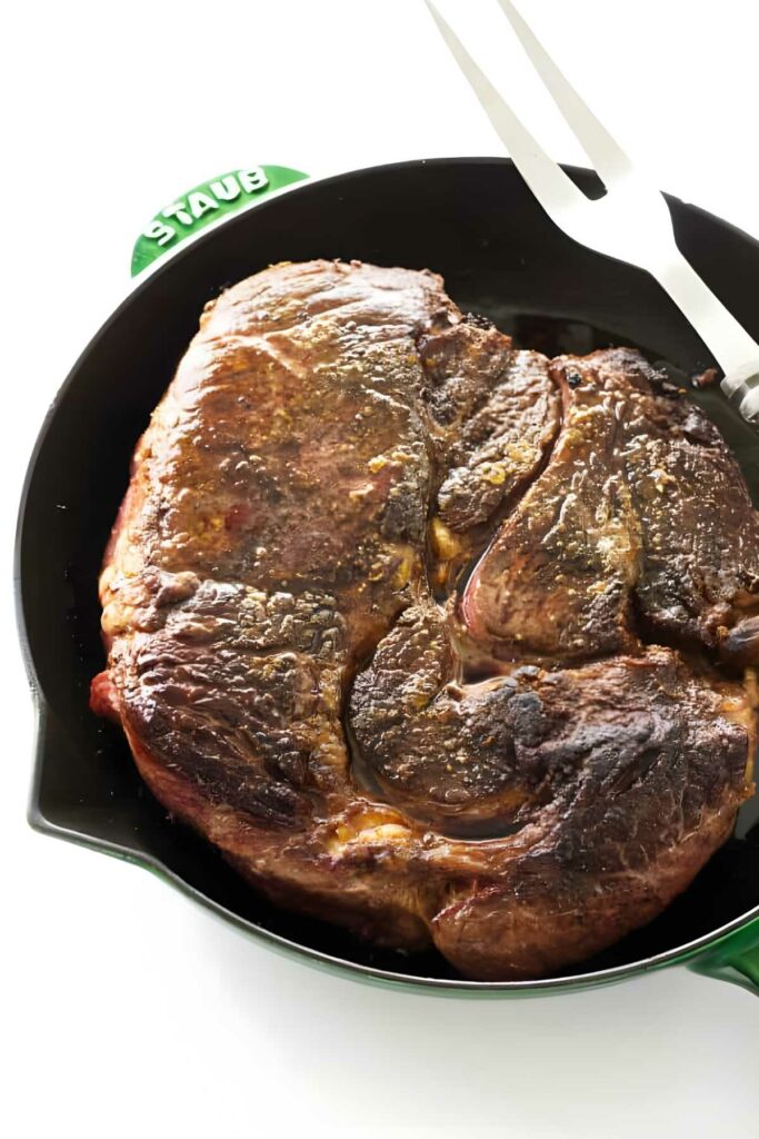 Seared beef roast in black skillet with metal fork, white background.
