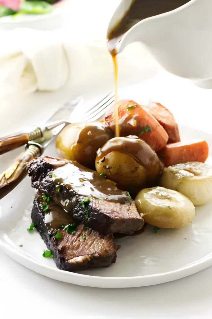 Pot roast with vegetables and gravy being poured, garnished with herbs.