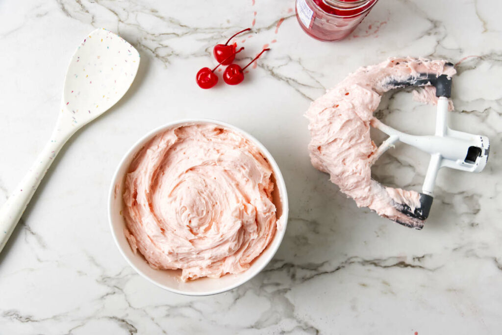 A bowl filled with cherry frosting and a beater next to the bowl.