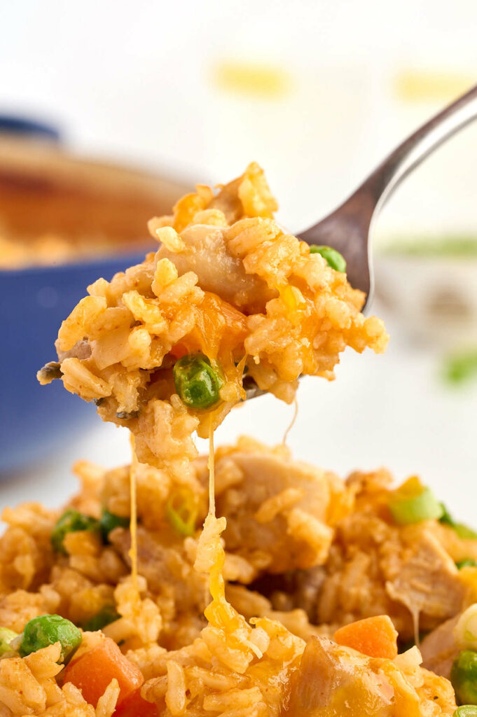 Fork lifts cheesy rice and chicken casserole with peas and carrots above a plate.