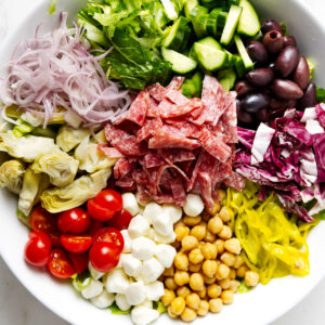 A mixed salad bowl with salami, cheeses, and vegetables on a white surface.