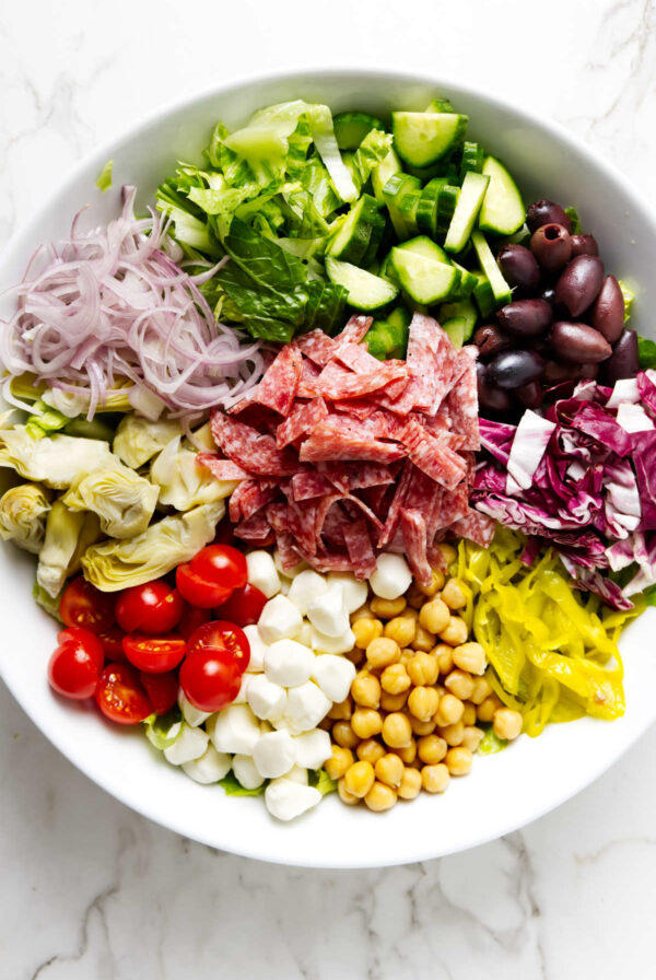 A mixed salad bowl with salami, cheeses, and vegetables on a white surface.