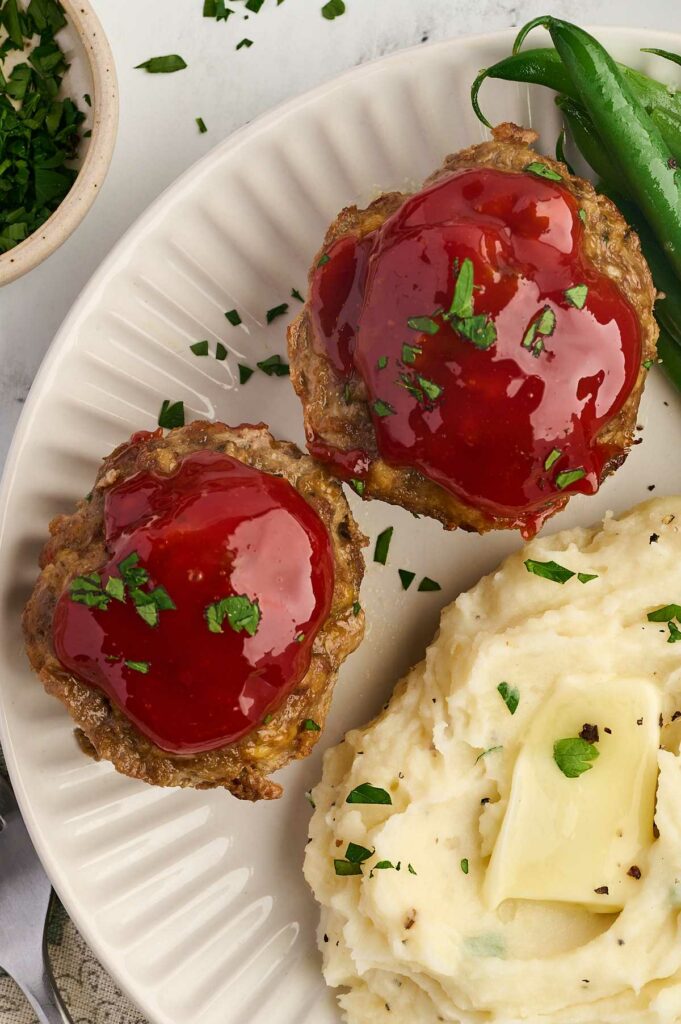 Two meatloaf muffin cups on a dinner plate with mashed potatoes and parsley.