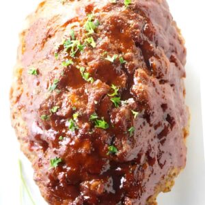 Glazed meatloaf with parsley garnish on a white surface.