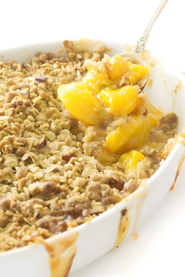 Peach crisp in white dish, spoon lifting portion, golden oat topping visible.