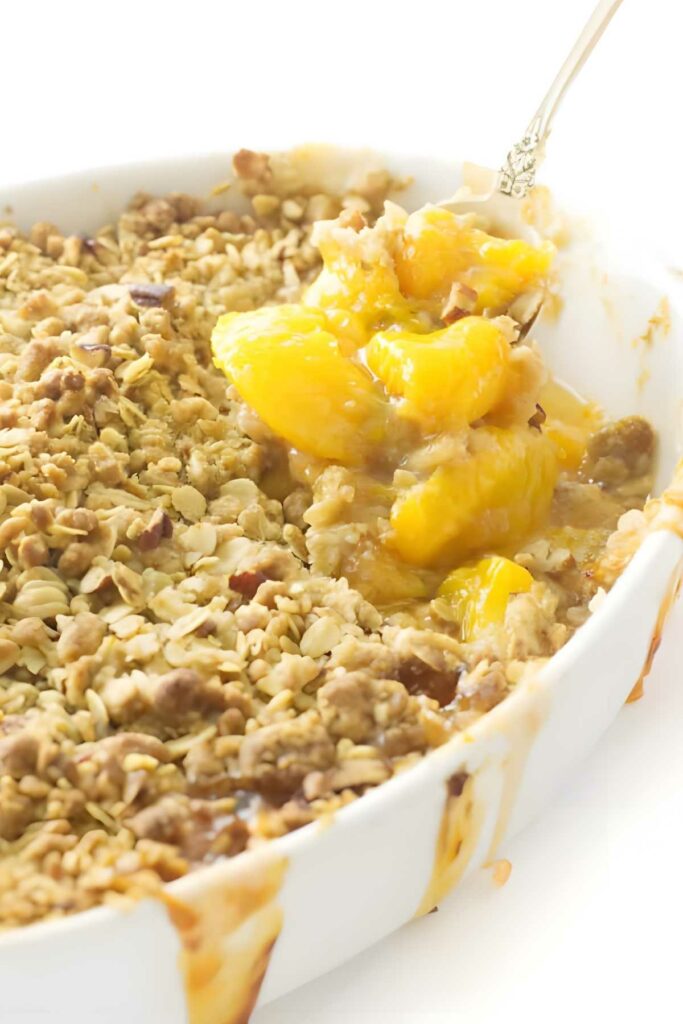 Peach crisp in a white dish with a spoon lifting a serving and golden oat topping visible.