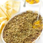 Baked peach crisp in white oval dish, serving on plate with fork nearby.