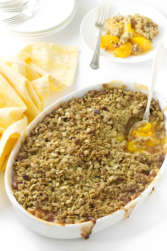Baked peach crisp in white oval dish, serving on plate with fork nearby.