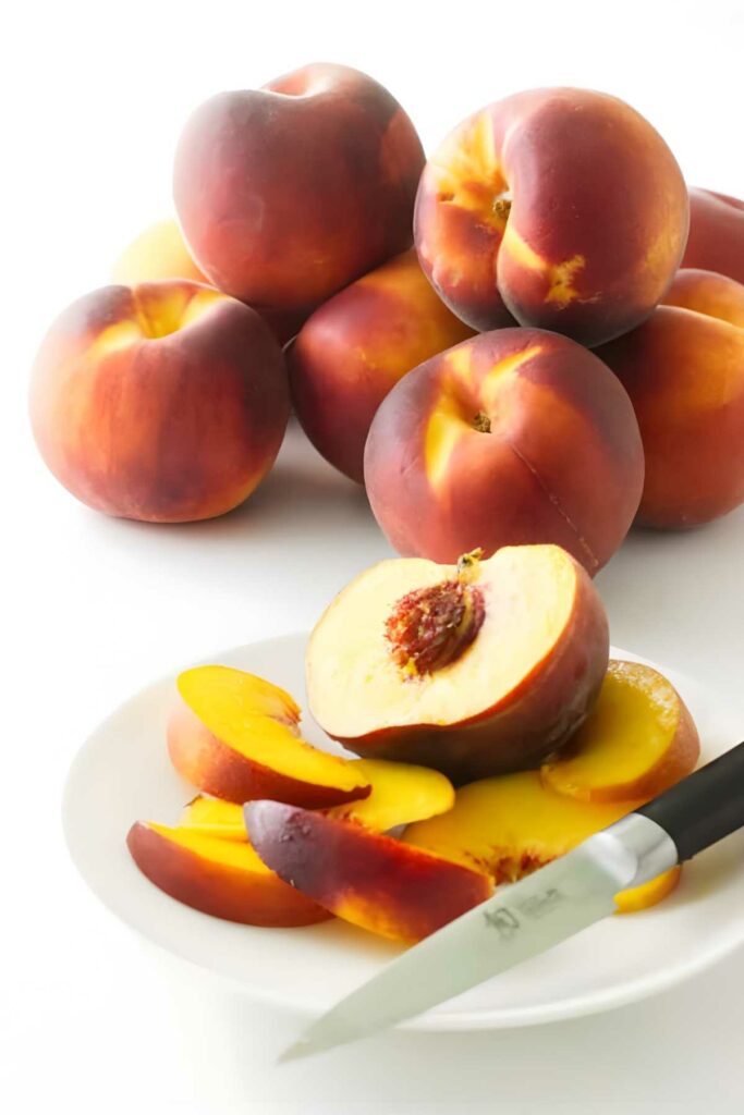 Sliced peach on a white plate with whole peaches in the background.