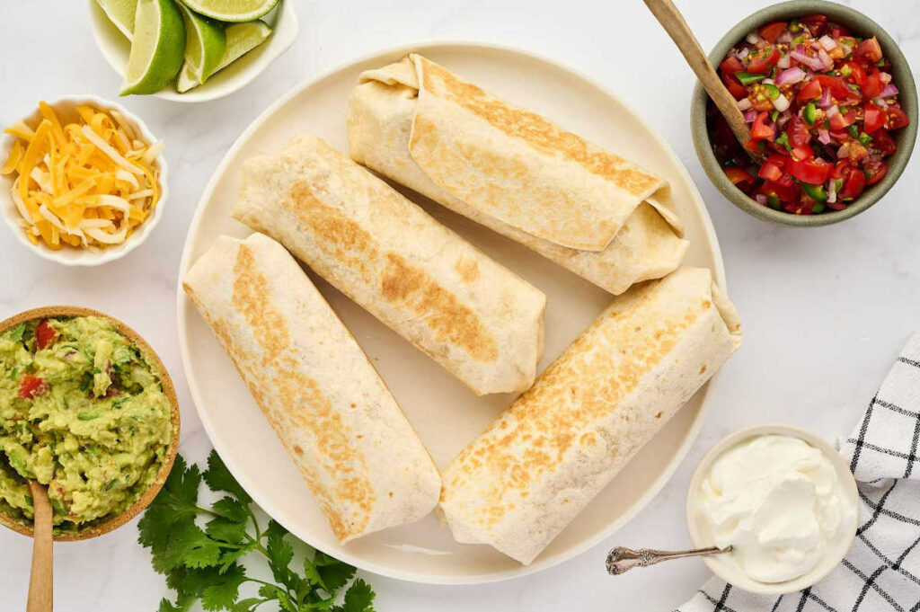 Four grilled chicken burritos on a plate with assorted toppings in small bowls nearby.