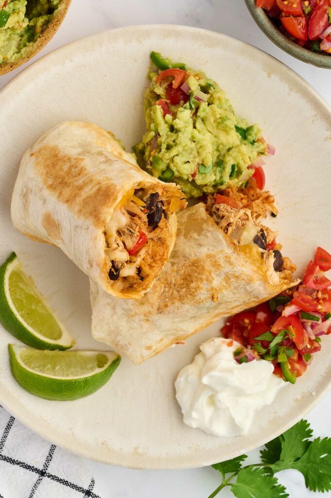 Two halves of a chicken and bean burrito on a plate with guacamole and salsa.