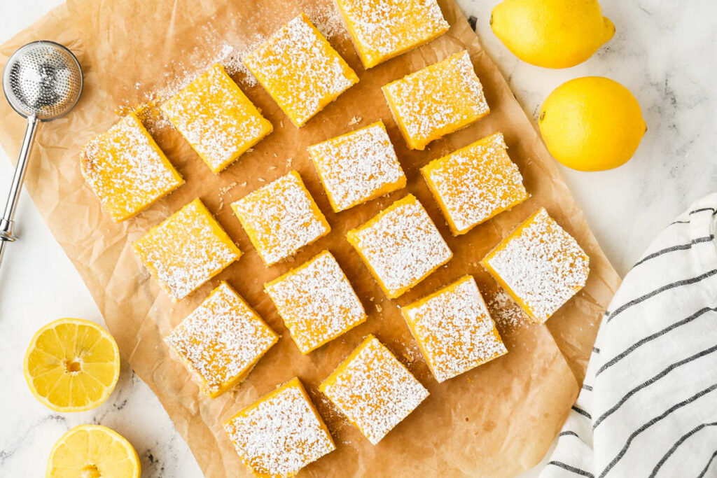 Twelve sliced lemon bars on parchment paper next to lemons.